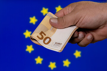 Hand holding a 50 euro banknote in front of a blue European Union flag with stars at a currency...