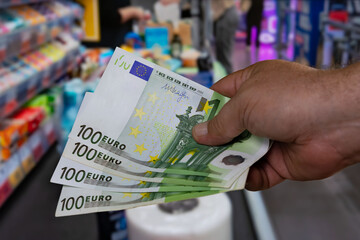 Hand holding several 100 euro banknotes in a supermarket checkout, with colorful product displays in the background during the day