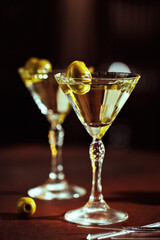 A pair of martini glasses with green olives, reflecting the warm glow of ambient lights on a wooden bar counter.