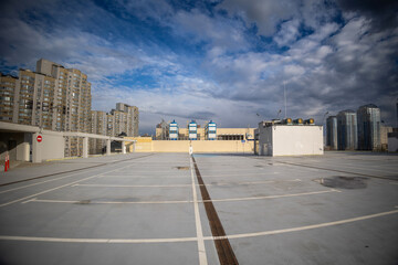 Wide-angle view of an empty rooftop parking lot with white marking lines. In the background, modern residential high-rise buildings of Kyiv's Osokorky district stand under a dramatic blue sky.