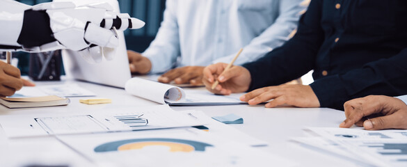 A diverse business team engaged in collaborative work with the assistance of a robotic arm, analyzing data and strategizing in a modern office setup.Noogenesis
