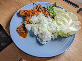 Traditional Indonesian meal with rice, grilled chicken satay, sambal, raw cabbage, and fresh basil leaves served on a blue plate