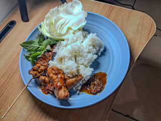 Traditional Indonesian meal with rice, grilled chicken satay, sambal, raw cabbage, and fresh basil leaves served on a blue plate