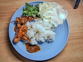 Traditional Indonesian meal with rice, grilled chicken satay, sambal, raw cabbage, and fresh basil leaves served on a blue plate