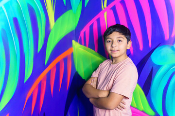 Young boy posing with crossed arms in front of vibrant neon graffiti wall