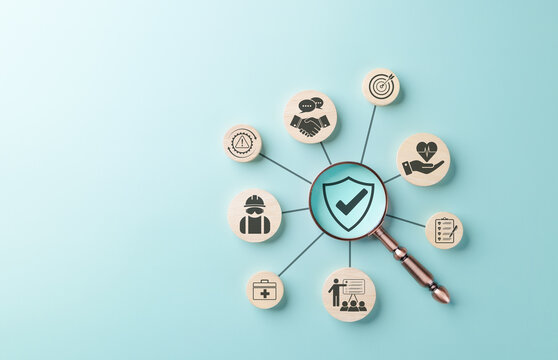 Work Safety and Compliance Concept. Wooden icons arranged in circle connected by lines, with magnifying glass focusing on shield symbol, representing workplace safety, compliance, and risk management.