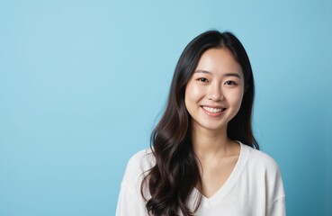 Fototapeta premium Happy young Asian woman smiles warmly in close-up. Dark-haired female in blue background. Young woman joyful expression and bright smile make her beautiful face stand out.