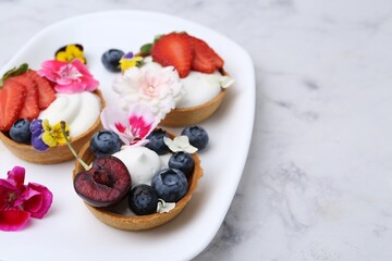 Sweet tartlets with berries and flowers on light marble table, closeup. Delicious dessert