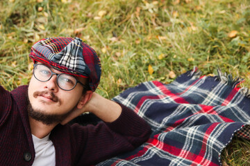 A young man in his 20s, with a beard and a checkered cap and scarf, is lying on the grass in autumn...