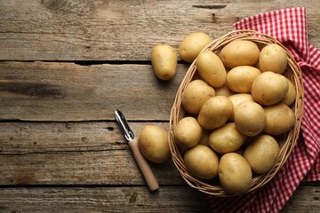 Fresh potatoes in basket and peeler on wooden table, flat lay. Space for text