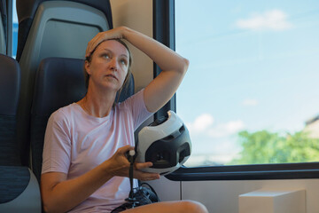 A woman sits on a train with bicycle helmet in her hands, looking thoughtful while gazing out the...