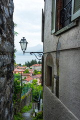 An old street lamp in a gap between old buildings overlooking houses with tiled roofs and Como Lake, Lombardia, Italy