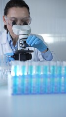 Female scientist wearing lab coat and protective blue gloves using microscope doing research in...