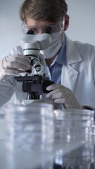 Man scientist wearing lab coat, white gloves, medical mask and protective glasses using microscope...