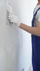 Close-up of man construction worker wearing gloves and blue overalls is using a putty knife to...
