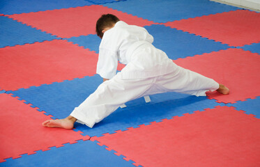 Young Kyokushin karate student performing a side split stretch on dojo mat, wearing a traditional white gi. Focus on flexibility, discipline, and physical preparation in Kyokushin karate training. © Olena Smerichka