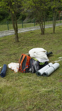 There are camping equipment, backpack, kmp mat, closed tent on the grass and trees in the background