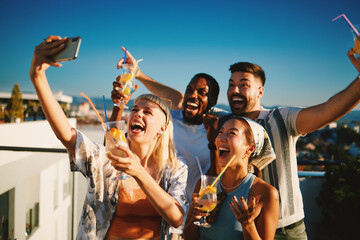 Group of young cheerful multi-ethnic friends taking selfie and enjoying drinks at sunset rooftop party