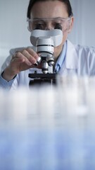 Woman scientist wearing lab coat and protective glasses is using microscope in modern laboratory...