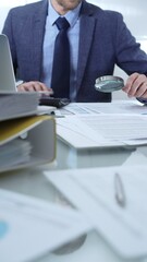 Businessman wearing blue suit is analyzing financial reports with magnifying glass and using...