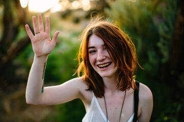 Young woman with red hair is joyfully waving while smiling in a lush outdoor environment filled with greenery, capturing a moment of happiness and connection with nature