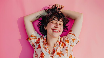 Young woman with curly hair is lying on a pink surface, smiling brightly with eyes closed, radiating joy and positivity, surrounded by a soft, colorful atmosphere that enhances her cheerful expression