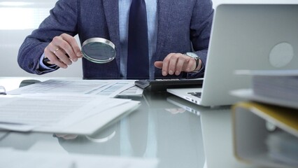 Businessman wearing blue suit is analyzing financial reports with magnifying glass and using...