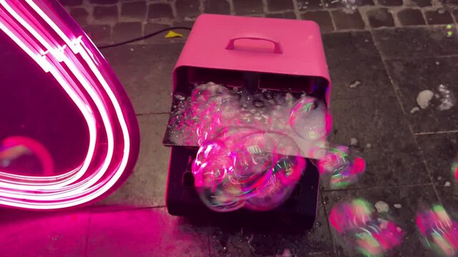 Iridescent soap bubbles emerging from pink bubble machine, floating near neon light against tiled floor, creating mesmerizing nightlife visual effect with vibrant, transparent spheres