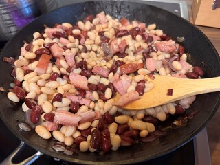 A hearty mix of red and white beans with crispy bacon and onions sizzling in a cast iron pan,...