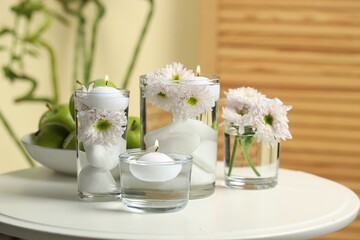 Burning candles and flowers in glasses of water on white table indoors