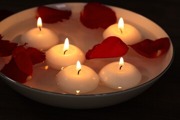 Burning candles and petals in bowl of water on table, closeup