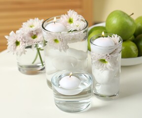 Burning candles and flowers in glasses of water on white table indoors, closeup