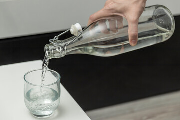 Hand pouring fresh water from glass bottle into tumbler on white table