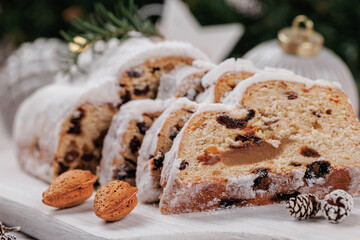 Traditional stollen bread sliced and covered with powdered sugar, presented on a white wooden board surrounded by festive ornaments, almonds, and pinecones.
