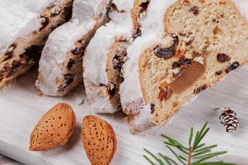 Close-up view of sliced Christmas stollen bread with dried fruits, almonds, and a powdered sugar coating, set on a white wooden board amidst festive decorations.