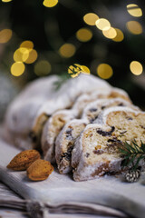 A traditional Christmas stollen sliced and presented on a wooden serving board surrounded by almonds, small pinecones, evergreen branches, and softly glowing holiday lights for a festive atmosphere.