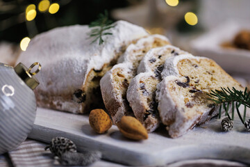 A rustic Christmas stollen sliced and served on a wooden board surrounded by almonds, pine branches, pinecones, and softly glowing holiday lights, creating a warm festive setting.