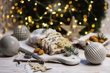 A festive Christmas stollen cut into slices, arranged on a rustic wooden board with almonds, pine sprigs, small pinecones, and twinkling golden holiday lights in a cozy and traditional winter setup.