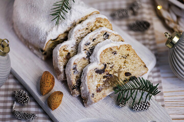 A close-up of a freshly sliced Christmas stollen with powdered sugar, set on a wooden board decorated with pine needles, almonds, pinecones, and blurred festive lights in the cozy background.