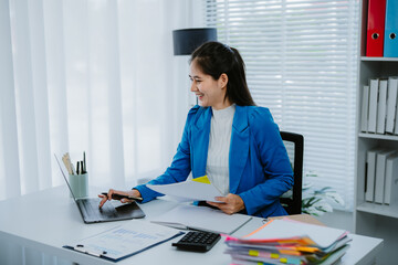 Young Asian woman working at home office, analyzing financial documents while using a laptop and...