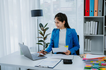 Young Asian woman working at home office, analyzing financial documents while using a laptop and...