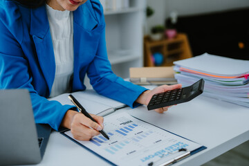 Young Asian woman working at home office, analyzing financial documents while using a laptop and...