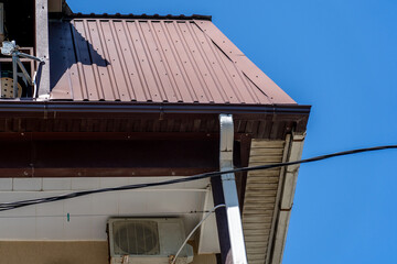 Close-up view of building's brown metal roof, matching gutters, white downspout, and outdoor air conditioning unit against clear blue sky