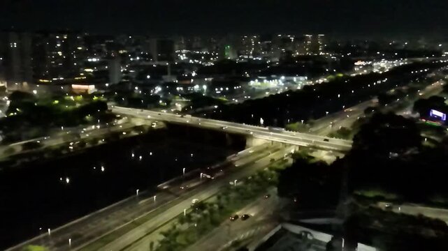 Nighttime aerial view of the Marginal Tiet&ecirc; highway in S&atilde;o Paulo, Brazil. Cars with lights on drive over a bridge crossing the Tiet&ecirc; River, with the illuminated city skyline in the background.