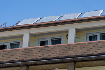 Multi-story building upper levels, featuring long row of angled solar thermal collectors mounted on brown tiled roof with yellow siding, white balcony columns and glass doors below