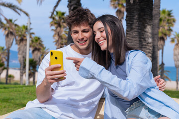 Young man and woman sit close on a wooden bench under palm trees by the sea, smiling as they look...