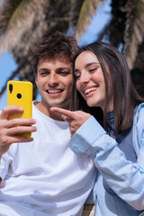 Young man and woman sit close on a wooden bench under palm trees by the sea, smiling as they look...