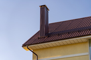 Architectural close-up of modern residential house with striking dark brown metal tile roof, matching chimney, and light yellow stucco wall under clear blue sky.
