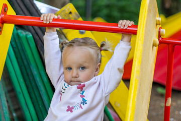 Fototapeta premium Blonde toddler girl with blue eyes and pigtails, dressed in pink, holds firmly onto bright red bar on colorful playground structure, showing determination.