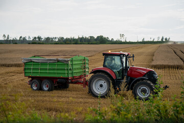 Obraz premium Modern red tractor with green trailer harvesting wheat in rural field during late summer, showcasing agricultural machinery, food production and farmland logistics in countryside